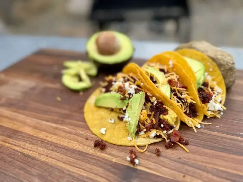 Cowboy Kent Rollins Bacon and Chorizo Breakfast Tacos, displayed on a wooden Mesquite cutting board. Corn tortillas, bacon, shredded cheese, queso fresco, and guacamole