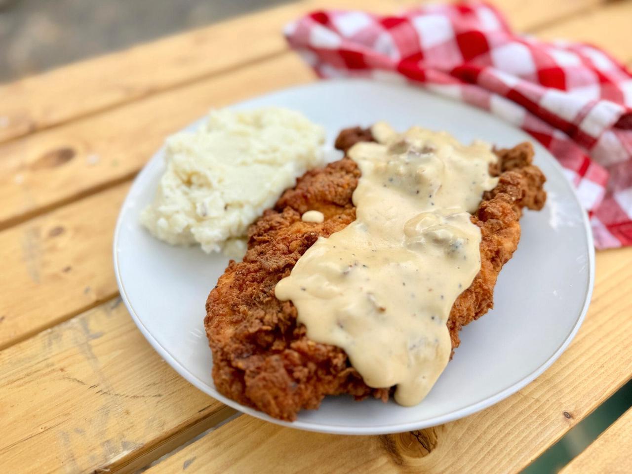 Cowboy Kent Rollins' Golden Crispy Chicken Fried Chicken with homemade gravy on a white plate, mashed potatoes on the side, with a red and white checked napkin in the background, on a picnic table.