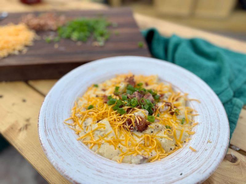 A bowl of loaded baked potato soup with heavy cream, cheddar cheese, bacon, and smoked sausage in a white bowl sits on a picnic table next to a green towel. In the background, green onions and shredded cheddar cheese sit on a dark cutting board.